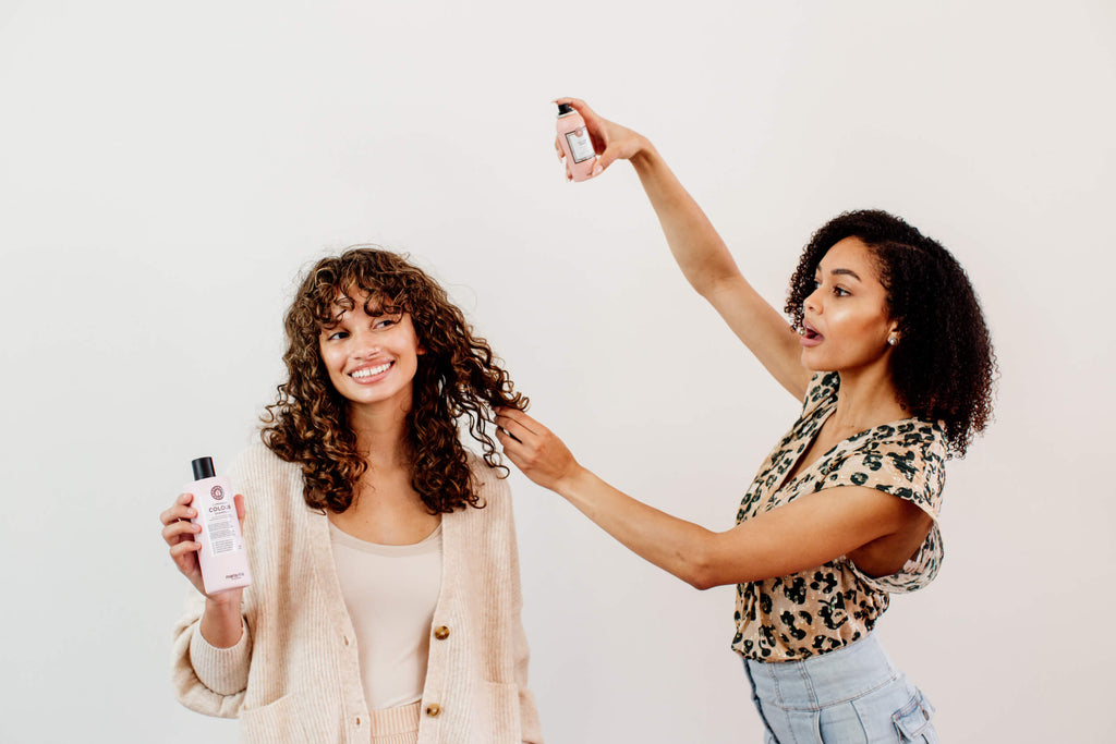 Two women with curly hair are doing each others hair. One one is spraying the other women's hair.
