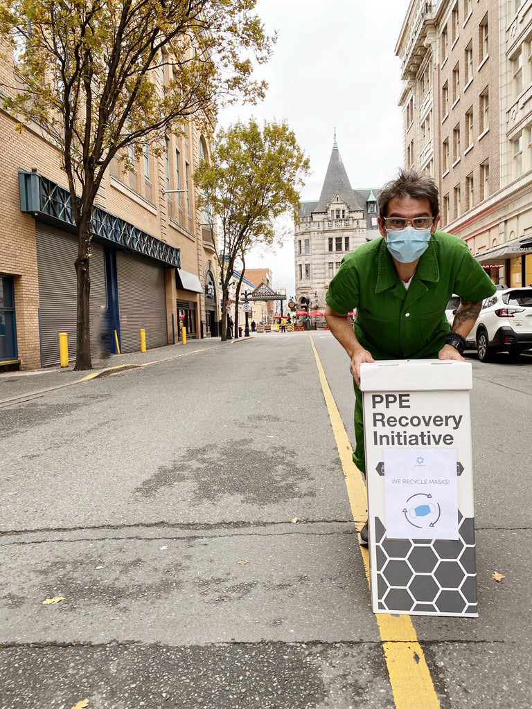 A man is standing in the middle of the street wearing a mask, with a sign saying PPE Recovery Initiative.
