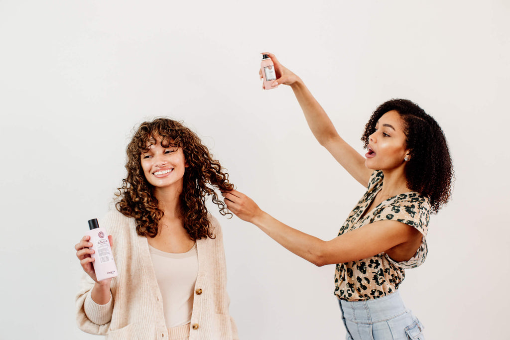 Two woman with curly hair are doing each others hair. One woman is hair spraying the other woman's hair.