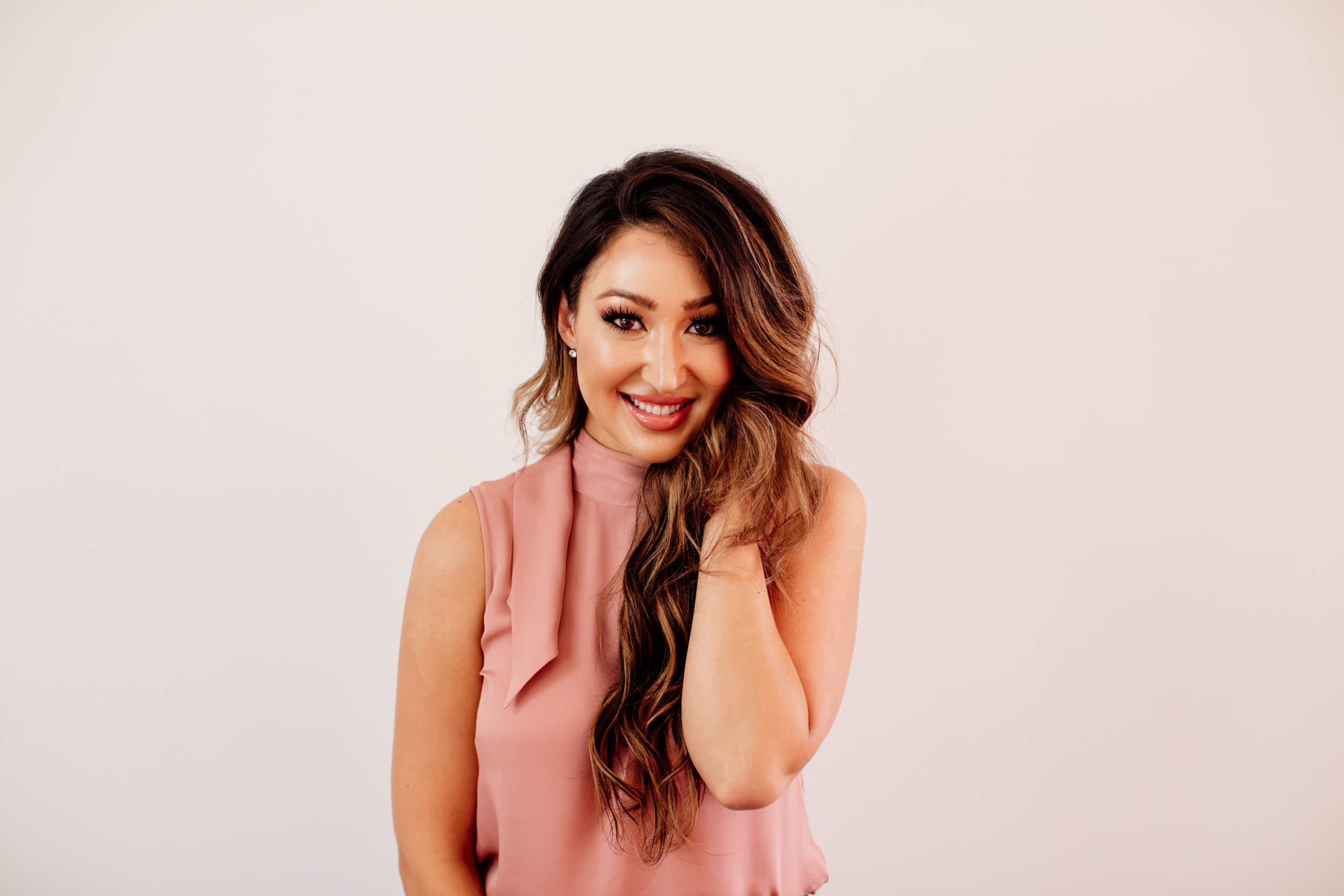 Woman wearing a pink top and dark long hair is smiling and holding her hair on one side. She has long hair extensions and newly highlighted hair.